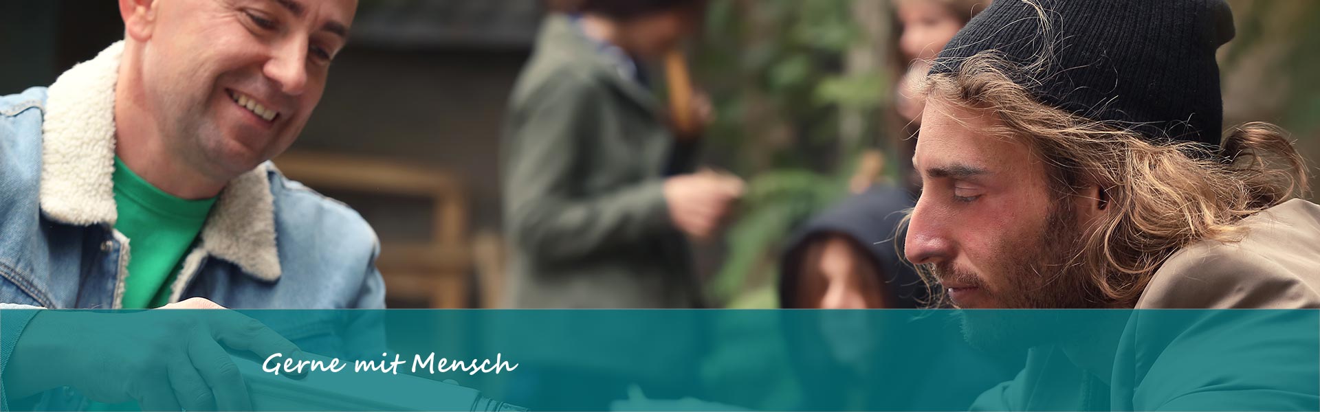 Ein Mann mit Jeansjacke schenkt einem jungen, wohnungslosen Mann mit Mütze Kaffee in eine Tasse ein. 3 weitere Personen sind verschwommen im Hintergrund zu sehen.